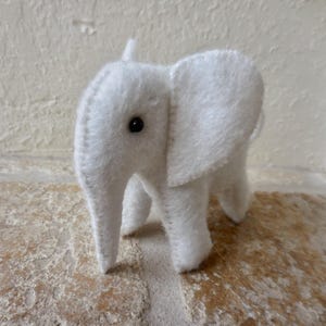 May include: A white plush elephant toy with a black button eye. The elephant is standing on a light brown surface.