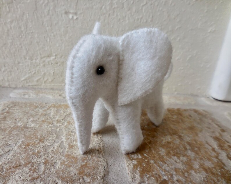 May include: A white plush elephant toy with a black button eye. The elephant is standing on a light brown surface.