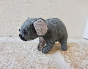 Può includere: Un giocattolo di peluche koala in feltro grigio e rosa. Il koala ha occhi neri e un naso nero.