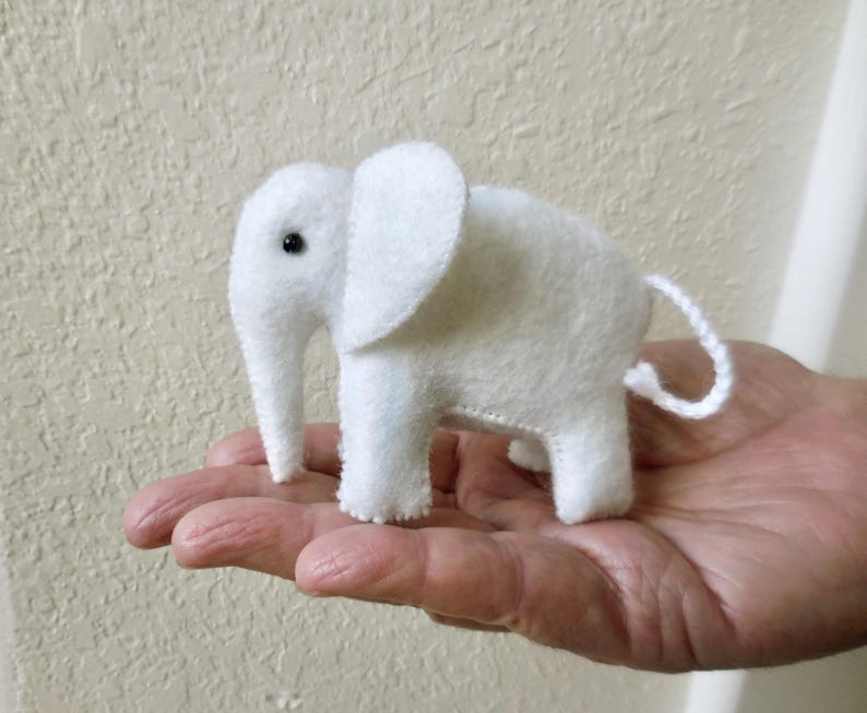 May include: A small white felt elephant toy with black eyes. The elephant is standing on a hand.