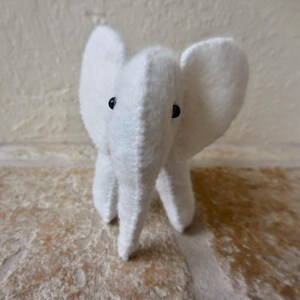 May include: A white plush elephant toy with black button eyes. The elephant is sitting on a light brown surface.