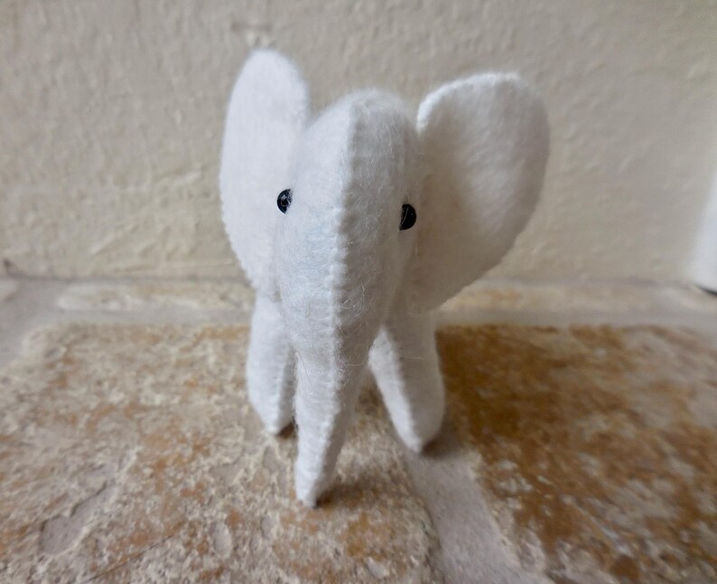 May include: A white plush elephant toy with black button eyes. The elephant is sitting on a light brown surface.