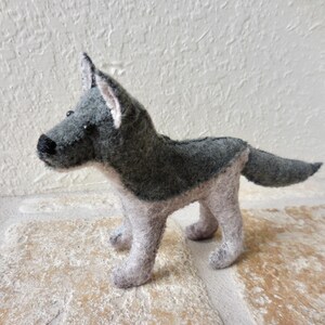 May include: A handmade felt dog figurine in shades of gray and white. The dog has a pointed snout, upright ears, and a fluffy tail. The toy is standing on a textured surface.