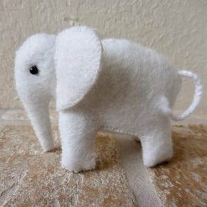 May include: A white felt elephant toy with black eyes. The elephant is standing on a light brown surface.