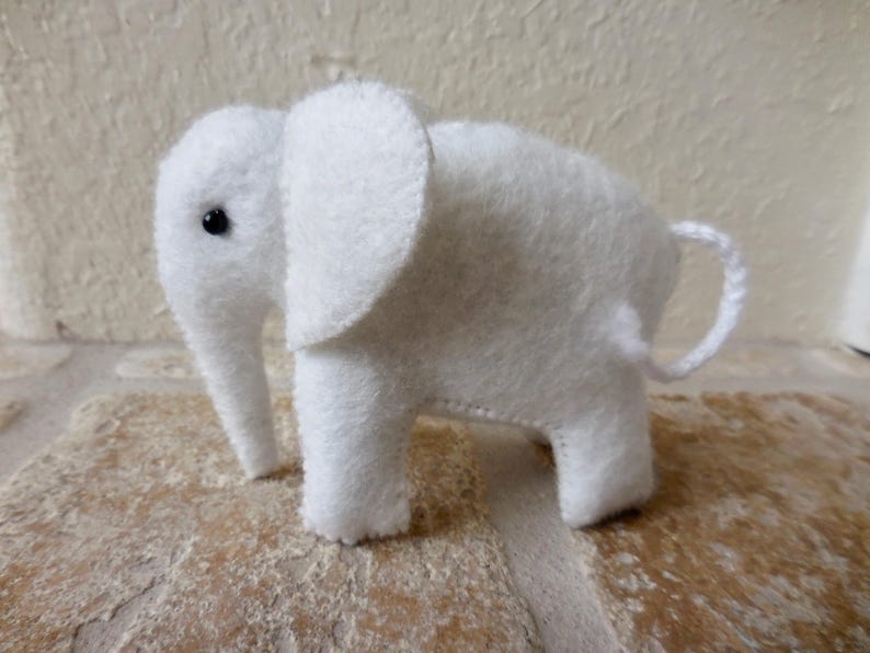 May include: A white felt elephant toy with black eyes. The elephant is standing on a light brown surface.