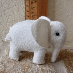 May include: A white felt elephant toy with a black button eye. The elephant is standing on a light brown surface.