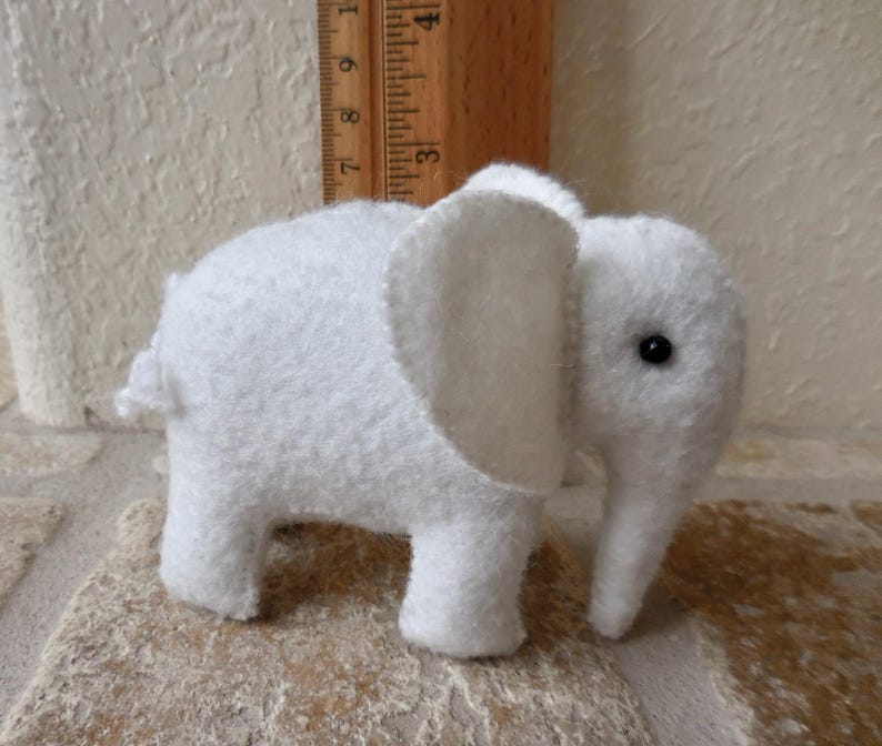 May include: A white felt elephant toy with a black button eye. The elephant is standing on a light brown surface.