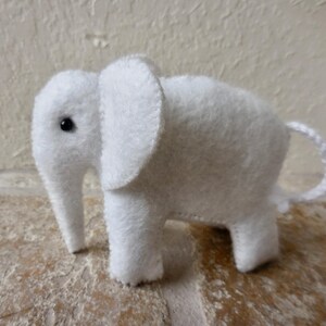 May include: A white felt elephant toy with a black button eye. The elephant has a long trunk and a small tail.