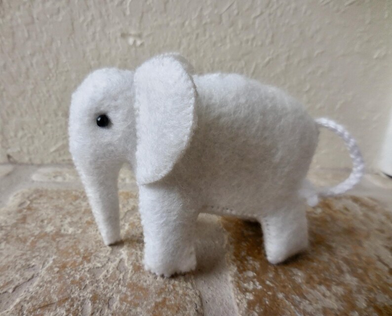 May include: A white felt elephant toy with a black button eye. The elephant has a long trunk and a small tail.