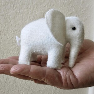 May include: A white felt elephant toy with black eyes. The elephant is sitting on a hand.