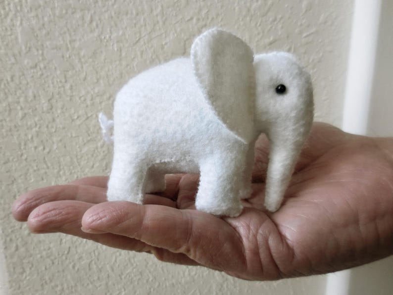 May include: A white felt elephant toy with black eyes. The elephant is sitting on a hand.
