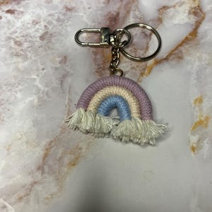 May include: A rainbow-coloured crocheted keychain with a gold chain and a silver key ring. The rainbow is made of pink, light blue, cream, and white yarn.