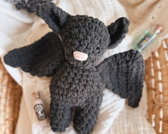 Puede incluir: Un murciélago de ganchillo gris oscuro hecho a mano con grandes alas, orejas y una nariz rosa. El murciélago es un artículo con temática de Halloween. En la imagen también hay botellas decorativas con corchos y un murciélago de fieltro negro.
