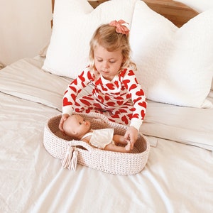 Può includere: Una bambina in pigiama con cuori rossi e bianchi tiene in mano un lettino per bambole in crochet con una bambola dentro. Il lettino è rosa chiaro e ha un fiocco bianco.
