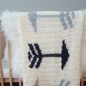 Puede incluir: Una manta de bebé de ganchillo blanco con un diseño de flecha en blanco y negro y flechas grises. La manta está drapeada sobre una cuna de madera.