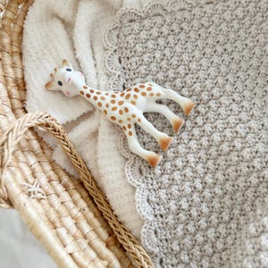 Pode incluir: Um cobertor de bebé em crochet bege com uma borda festonada está numa cesta de vime com um cobertor de pelúcia branco e um mordedor em forma de girafa.