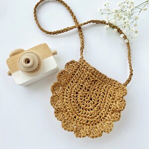 May include: A small, brown, crocheted bag with a scalloped edge and a long brown strap. The bag is on a white surface with a wooden toy camera.