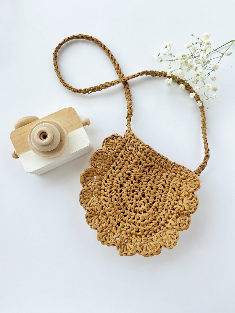 May include: A small, brown, crocheted bag with a scalloped edge and a long brown strap. The bag is on a white surface with a wooden toy camera.