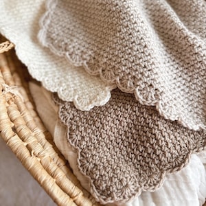 May include: A close-up shot of three crocheted blankets in cream and tan, each with a scalloped edge. The blankets are folded and nested inside a woven basket, creating a cosy and textured display. The soft, neutral colours suggest a handmade, comforting item.