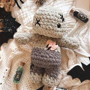 May include: A handmade crochet zombie doll in shades of gray and purple, with black cross-stitch eyes and mouth. The doll has a pink heart-shaped detail and is surrounded by Halloween decorations, including bats and potion bottles.