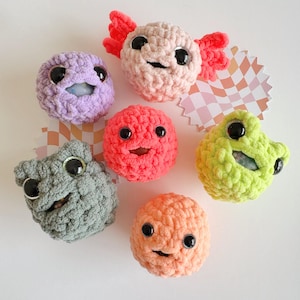 May include: Six crocheted plush toys with smiling faces. The toys are in various colors, including pink, purple, orange, green, and gray. The toys are all about 2 inches in diameter.