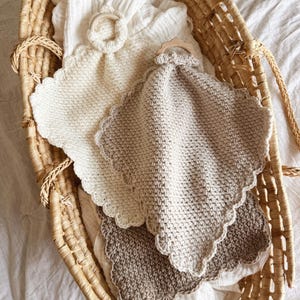 May include: A woven basket holds several baby comfort blankets. The blankets are in shades of cream, beige, and brown, with scalloped edges. One blanket has a wooden ring attached. The basket is lined with a white muslin cloth.