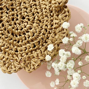 May include: A close-up of a woven straw coaster with a scalloped edge. The coaster is on a pink surface with white baby's breath flowers.