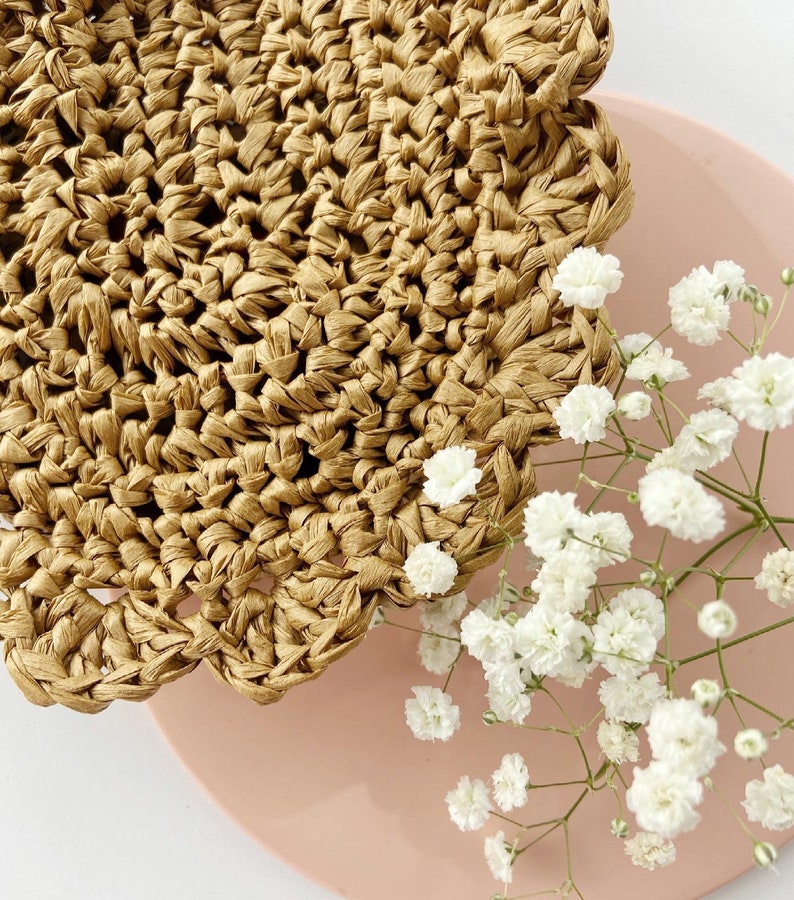 May include: A close-up of a woven straw coaster with a scalloped edge. The coaster is on a pink surface with white baby's breath flowers.