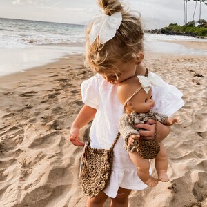 May include: A young girl in a white dress holds a doll with a brown knitted sweater and a white bow. The girl is wearing a brown crocheted bag with a white bow in her hair. The girl and doll are standing on a sandy beach.