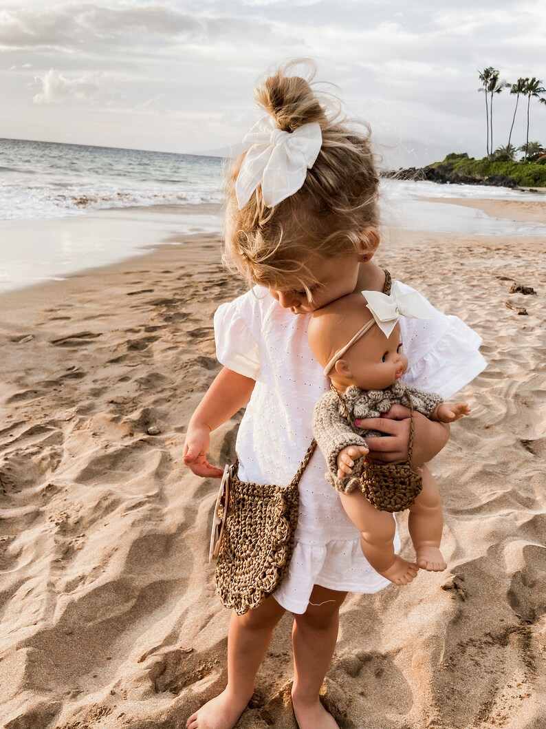 May include: A young girl in a white dress holds a doll with a brown knitted sweater and a white bow. The girl is wearing a brown crocheted bag with a white bow in her hair. The girl and doll are standing on a sandy beach.