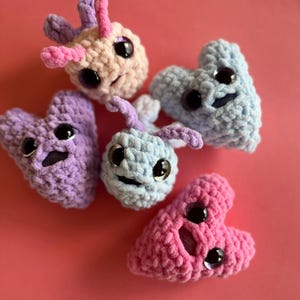 May include: A collection of five handmade crochet creatures in various pastel colors, including pink, purple, and blue. Each creature has large, shiny black eyes and a smiling face. The plush toys are arranged on a solid pink background.