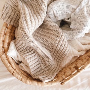 May include: A woven wicker basket containing a beige ribbed knit blanket, a white muslin cloth, and a white long-sleeved garment featuring a fine black dot pattern with a vertical row of snap buttons along the front side of the clothing item.