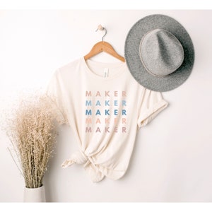 Op de afbeelding: Een beige T-shirt met het woord "MAKER" vijf keer herhaald in een gestapelde vorm. Elk woord is in een andere tint blauw, roze en perzik.