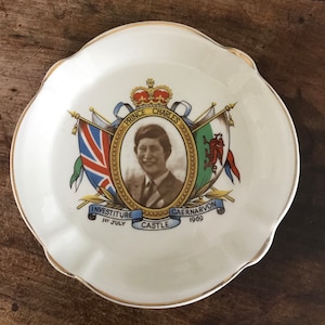 Peut inclure: Une assiette en céramique blanche avec un bord doré présente une image commémorative du prince Charles. L'image comprend un portrait du prince, une couronne et des drapeaux représentant l'Angleterre, l'Écosse et le Pays de Galles. Le texte "Investiture 1er juillet Château de Caernarvon 1969" est imprimé sous l'image.