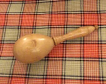 May include: A light-colored wooden maraca with a rounded body and a long handle. The maraca is made of a light-colored wood and has a smooth finish. The background is a plaid pattern in red, green, and black.