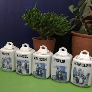 Könnte beinhalten: Fünf weiße Keramik-Gewürzgläser mit blau-weißen Designs. Die Gläser sind mit "peper", "kaneel", "kruiden", "foelie" und "naglen" beschriftet.