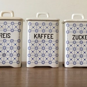 Könnte beinhalten: Drei weiße Keramikbehälter mit Deckeln und Griffen. Jeder Behälter hat ein blaues Blumenmuster und ein schwarzes Etikett mit der Aufschrift "REIS", "KAFFEE" und "ZUCKER". Die Behälter stehen auf einer Holzoberfläche.