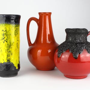 May include: Three ceramic vases in a studio setting. The vase on the left is tall and cylindrical, with a yellow and black color scheme. The middle vase is orange with a handle. The vase on the right is red with a black textured top.