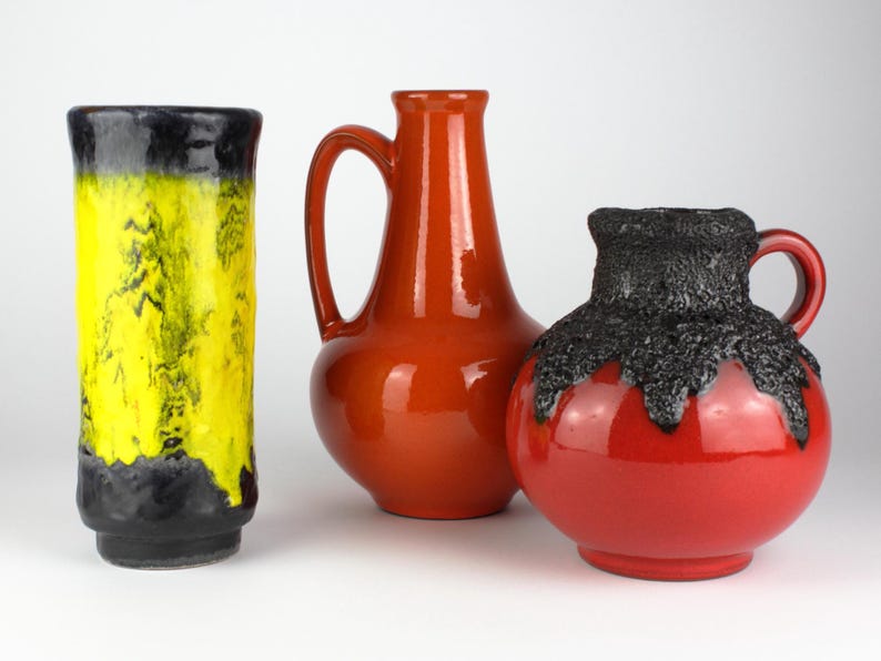 May include: Three ceramic vases in a studio setting. The vase on the left is tall and cylindrical, with a yellow and black color scheme. The middle vase is orange with a handle. The vase on the right is red with a black textured top.