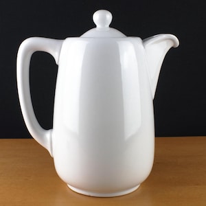 Op de afbeelding: Een witte keramische koffiepot met een gladde, glanzende afwerking, voorzien van een hoog cilindrisch lichaam, een gebogen handvat aan de linkerkant, een schenktuit aan de rechterkant en een afneembaar deksel met een kleine ronde knop.