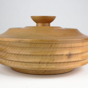 Puede incluir: Un tazón de madera con tapa, que presenta un diseño único y en espiral. El tazón está hecho de madera marrón claro y tiene un acabado liso y pulido.