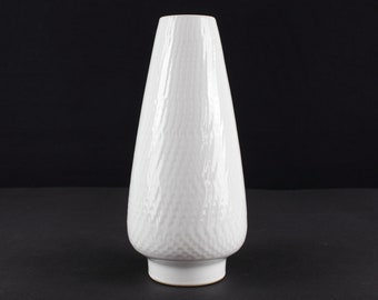 May include: A white ceramic vase with a textured surface. The vase is tall and narrow, with a rounded base and a slightly flared opening.
