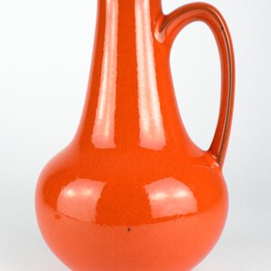 May include: A glossy, orange ceramic pitcher with a curved handle. The pitcher has a bulbous base that tapers to a narrow neck and a flared rim. The handle is a darker shade of orange. The pitcher is likely used for serving beverages.