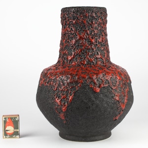 May include: A large, textured ceramic vase with a black base and a red, lava-like top. The vase has a bulbous body and a narrow neck. A matchbox is next to the vase.