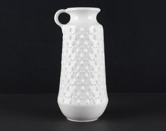 May include: White ceramic vase with a textured pattern and a handle. The vase is tall and slender.