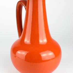 May include: A glossy, orange ceramic pitcher with a rounded base and a slender neck. The pitcher has a curved handle and a dark brown interior. The overall design is simple and elegant, suitable for serving beverages or as a decorative item.