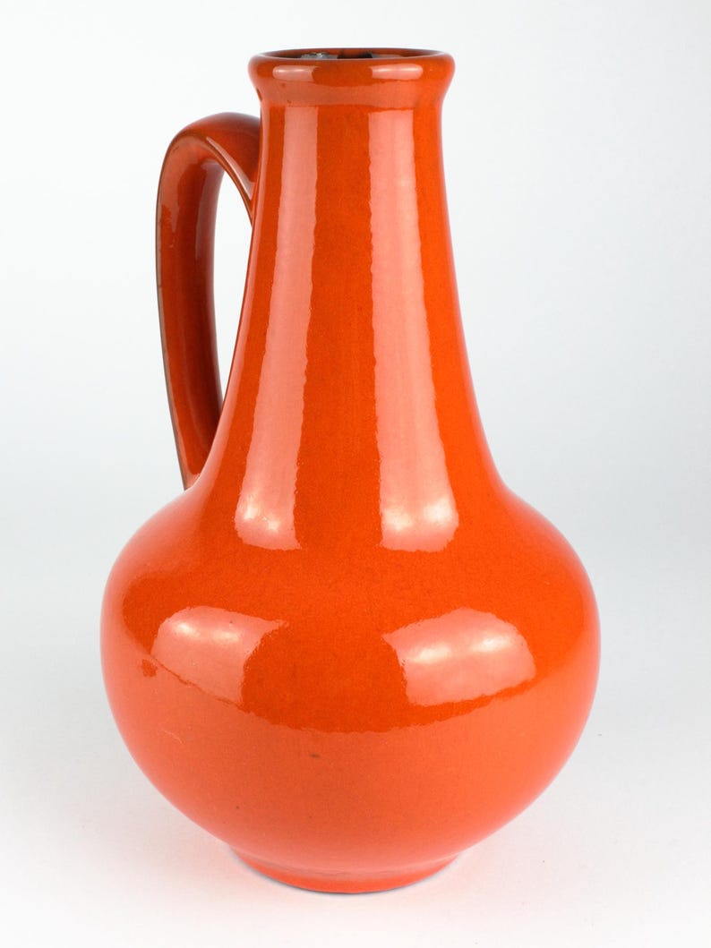 May include: A glossy, orange ceramic pitcher with a rounded base and a slender neck. The pitcher has a curved handle and a dark brown interior. The overall design is simple and elegant, suitable for serving beverages or as a decorative item.