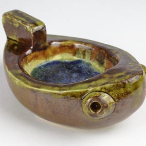 Puede incluir: Un cuenco de cerámica con un esmalte verde, marrón y azul. El cuenco tiene una forma única, que recuerda a un pez con un pico en el lateral.