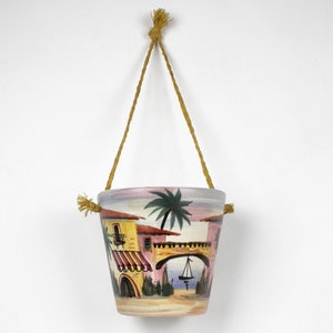 May include: A ceramic hanging planter featuring a painted scene of a pink building with a yellow awning, a palm tree, and a bridge over water. The planter is suspended by a brown rope.