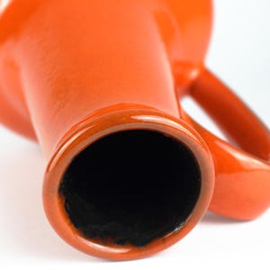 May include: Close-up of a vibrant orange ceramic vase with a glossy finish. The vase has a wide opening and a handle. The interior is black. The design is simple and modern, suitable for home decor.